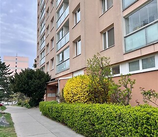 Venkovní pohled na bytový dům s moderní fasádou. V popředí dobře udržovaný keř a chodník, vhodný pro pohodlný přístup k budově.