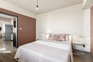 Prostorná ložnice s dřevěnou podlahou, moderními lampami a světlými stěnami, doplněná velkou postelí se stylovými polštáři.