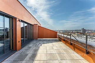 Prostorná terasa s dlažbou, moderní zábradlí a velká okna nabízejí panoramatický výhled na město.