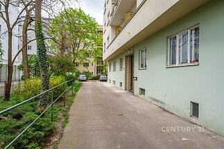 Venkovní prostor bytového domu s asfaltovou příjezdovou cestou a zelení. K dispozici jsou venkovní parkovací místa a udržovaná zahrada.