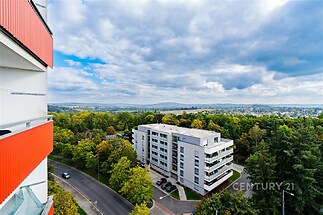 Pohled z balkonu nabízí panoramatický výhled na moderní bytový dům obklopený zelení, ideální pro klidné bydlení s dobrou dostupností.