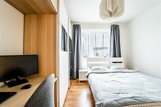 Světlá ložnice s parketovou podlahou, velká okna pro přirozené osvětlení, moderní vestavěná skříň a stylová závěsná lampa.