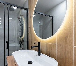 Moderní koupelna, dřevěný obklad, skleněný sprchový kout, oválné zrcadlo s LED osvětlením, elegantní černé vodovodní baterie
