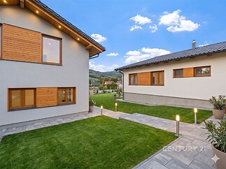 Dvorek s moderní zámkovou dlažbou, udržovaným trávníkem a venkovním osvětlením, obklopený domy s dřevěnými okenicemi a novou fasádou.