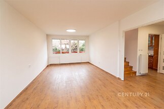 Světlý obývací pokoj s dřevěnou podlahou, velká okna s výhledem do zahrady, moderní bílá stěna, prostorný a útulný interiér.