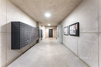 Chodba s betonovými stěnami, moderními poštovními schránkami a dlažbou, nabízí praktický a světlý prostor v rezidenční budově.