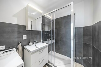 Moderní koupelna s prostorným sprchovým koutem, šedými obklady a elegantním nábytkem. Velké zrcadlo a čistý design vytvářejí světlý prostor.