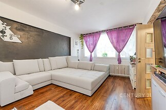 Světlý obývací pokoj s moderní rohovou sedačkou, parketová podlaha, velká okna s fialovými závěsy, útulný a svěží prostor.