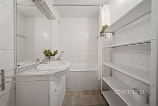 Kompaktní koupelna s dlažbou a bílými obklady, vybavená vanou a umyvadlem s úložnou skříňkou, jednoduchý a čistý design.