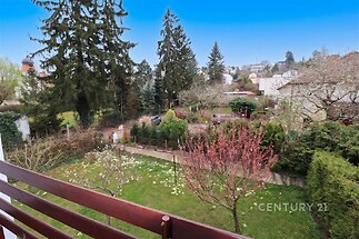 Výhled z balkonu na zahradu s ovocnými stromy, vzrostlé jehličnany a udržované keře, poskytující poklidné a zelené prostředí v rezidenční čtvrti.