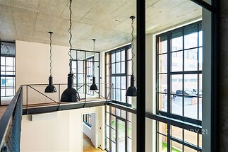 Moderní interiér s vysokým stropem, velká okna a parketová podlaha, industriální styl s kovovými prvky, ideální pro loftové bydlení.