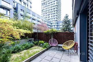 Malá terasa s dlažbou, oplocením a moderním venkovním nábytkem. Obklopena zelení a s výhledem na městskou architekturu.