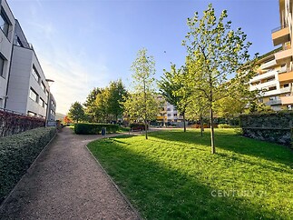 Moderní rezidenční zahrada s udržovanou zelení, cestičkou z dlažby a lavičkami, obklopená moderními bytovými domy.
