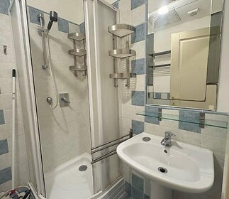 Koupelna s moderním sprchovým koutem, světlé dlaždičky a elegantní umyvadlo, dekorativní dlažba na podlaze a praktické poličky na stěnách.