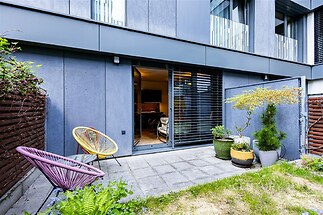 Terasa s moderními dlaždicemi, obklopená zelení, s pohodlným venkovním posezením a velkými prosklenými dveřmi vedoucími do interiéru