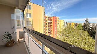 Zasklený balkón s dlažbou nabízí výhled na barevné panelové domy a zeleň, ideální pro relaxaci a moderní bydlení ve městě.