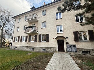 Fasáda bytového domu s balkonem a zatažitelnými okenicemi, travnatá předzahrádka, klidná lokalita, třípodlažní budova s renovovanou střechou.