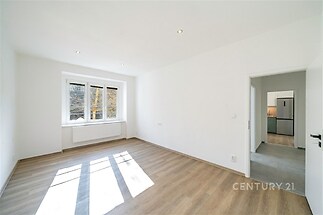 Světlý obývací pokoj s parketovou podlahou, velká okna poskytující dostatek přirozeného světla, moderní vzhled a přímý vstup do kuchyně.