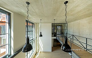 Industriální obývací pokoj s betonovým stropem, velkými okny a dřevěnou podlahou, moderní loft styl a kovové zábradlí.