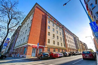 Historická budova s červenou cihlovou fasádou a velkými okny, nacházející se v městské části s parkovacími místy.