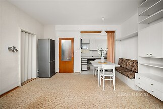 Jídelní kout s vestavěnými skříněmi, kobercovou podlahou a bílým nábytkem, propojený s malou kuchyní, působí útulně a funkčně.