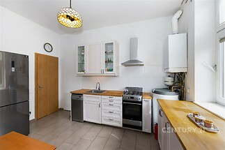 Kuchyň s dlažbou a dřevěnou pracovní deskou, moderní vybavení včetně plynového sporáku a vestavěných skříní, světlý interiér.