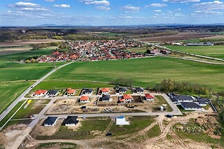 Výhled na nově budovanou rezidenční čtvrť s moderními rodinnými domy, obklopenou zelenými poli a blízko malebné vesnice v klidné lokalitě.