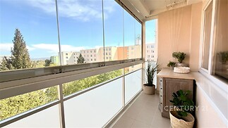 Zasklená lodžie s dlažbou, moderním nábytkem a výhledem na okolní zástavbu. Prosvětlený prostor s dekorativními rostlinami.