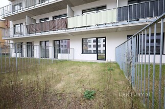 Přízemní terasa s trávníkem a moderní zábradlím, přiléhající k rekonstruovanému bytovému domu, s velkými okny a balkony.