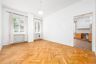 Světlý obývací pokoj s parketovou podlahou a velkými okny přechází do moderní kuchyně.