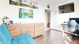 Světlý obývací pokoj s dřevěnou podlahou, moderním nábytkem a velkou televizí, ideální pro pohodlný odpočinek a práci z domova.