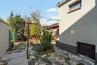 Zahrada s přírodní kamennou dlažbou, obklopená zelení, nabízí klidný prostor k relaxaci. Soukromý dům se světlou omítkou a jednoduchým designem.