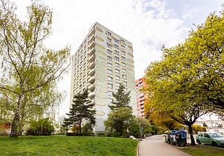 Venkovní pohled na vysokopodlažní obytný dům s balkonky, obklopený zelení a upraveným parkem, ideální pro městské bydlení.