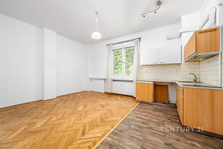 Světlý obývací pokoj s parketovou podlahou, moderní kuchyňskou linkou a velkým oknem, dřevěné detaily v kombinaci s dlažbou, nabízí útulný prostor.