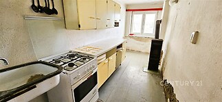 Kuchyň s vintage dřevěnými skříňkami, sporákem a velkým oknem, vyžaduje rekonstrukci, dobře prosvětlena denním světlem.