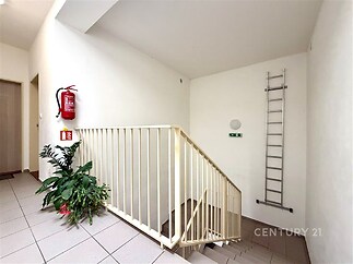 Chodba se schodištěm, světlá výmalba, keramická dlažba, nouzové osvětlení a bezpečnostní prvky, čisté a udržované prostředí.