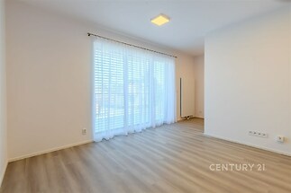 Světlý obývací pokoj s parketovou podlahou, velká okna s bílými závěsy, moderní design a prostor pro relaxaci.