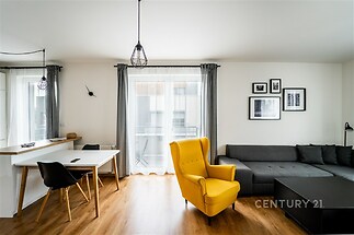 Světlý obývací pokoj s parketovou podlahou, velkými okny a moderním nábytkem, včetně žlutého křesla a šedé pohovky.