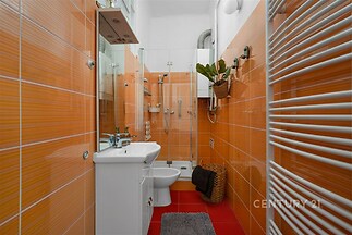 Moderní koupelna s oranžovými dlaždicemi, prosklený sprchový kout, bílé sanitární vybavení, topný žebřík, světlé a dobře udržované.