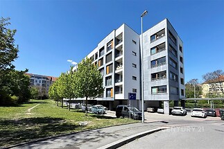 Moderní bytový dům s balkony v klidné lokalitě, fasáda s čistými liniemi, parkoviště vedle budovy, obklopený zelenými plochami a stromy.