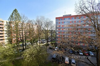 Výhled z bytu na sídlišti s panelovými domy, stromy a parkovištěm, klidná lokalita s dostupností městské infrastruktury