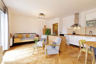 Světlý obývací pokoj s parketovou podlahou a moderní kuchyní, nabízí velká okna a otevřený prostor ideální pro pohodlné bydlení.