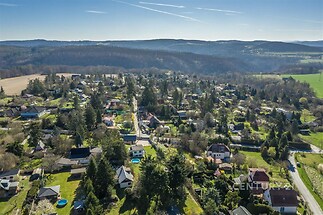 Letecký pohled na malebnou vesnici s rodinnými domy, upravené zahrady a okolní lesy. Klidná lokalita, ideální pro bydlení v přírodě.