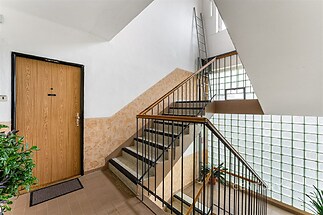 Světlá vstupní hala s dlažbou, dřevěnými dveřmi a velkými okny propouštějícími přirozené světlo, v udržovaném bytovém domě