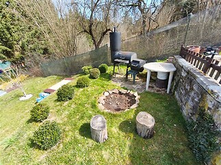 Zahradní prostor se zahradním grilem, dřevěnými špalky a kulatým stolem, obklopený udržovaným trávníkem a zelení, ideální pro relaxaci.