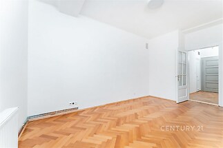 Světlý obývací pokoj s parketovou podlahou a bílými stěnami, moderní dveře vedou do další místnosti, celkově moderní styl.