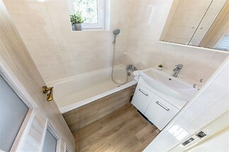 Kompaktní koupelna s parketovou podlahou, světlé dlažby, moderním umyvadlem a velkým zrcadlem, přirozeně osvětlená oknem.