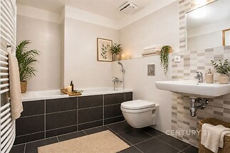 Moderní koupelna s dlažbou, světlými obklady a velkou vanou. Místnost zdobí dekorativní rostliny a elegantní zrcadlo.