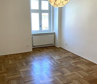 Světlý obývací pokoj s parketovou podlahou, velkým oknem a moderním osvětlením, ideální pro rekonstrukci či moderní úpravu prostoru.