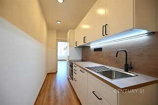 Moderní kuchyň s dřevěnou podlahou, bílými skříňkami a vestavěným osvětlením. Úzký prostor nabízí praktický a stylový design.